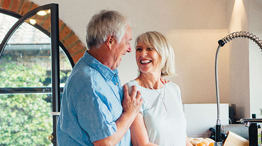 A couple smiling at each other, stood in the kitchen with patio doors in the background