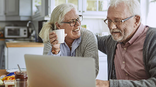 Lady holding a cup, smiling at her partner who is looking at a laptop in the kitchen