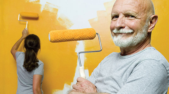 A man holding a roller smiling at the camera, a woman painting the wall yellow with a roller in the background facing away